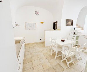 Un intérieur aux murs blancs, un sol carrelé, une table blanche avec des chaises, un petit bol jaune, un tableau encadré et une horloge au mur.