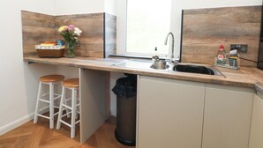 Una cocina con ventana, un lavamanos, taburetes, un cubo de basura y un reloj en la pared.