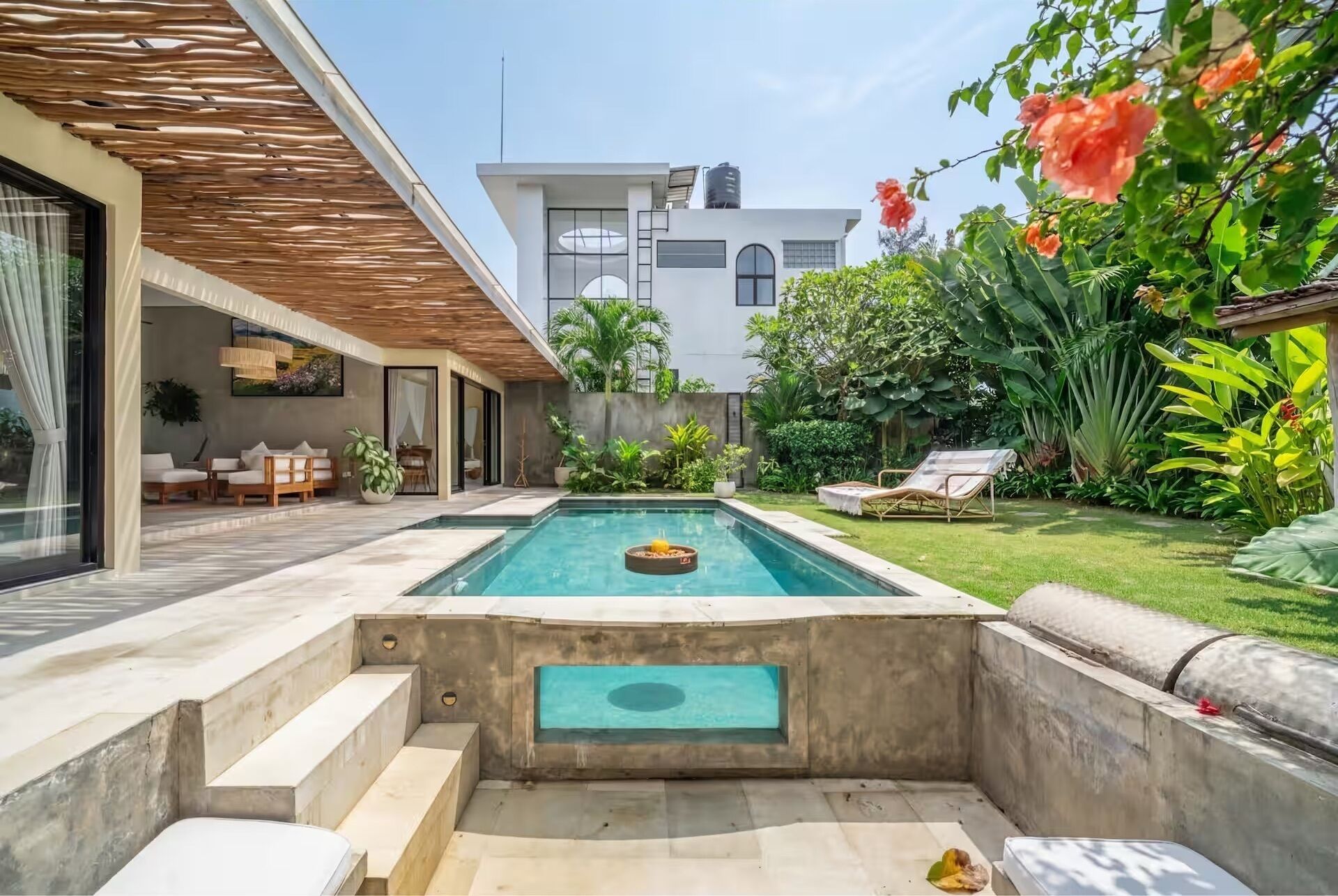A modern outdoor pool area with a built-in hot tub, a seating area with a table and chairs, and lush greenery.