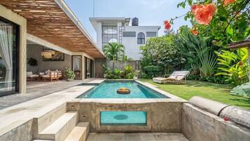 A modern outdoor pool area with a built-in hot tub, a seating area with a table and chairs, and lush greenery.