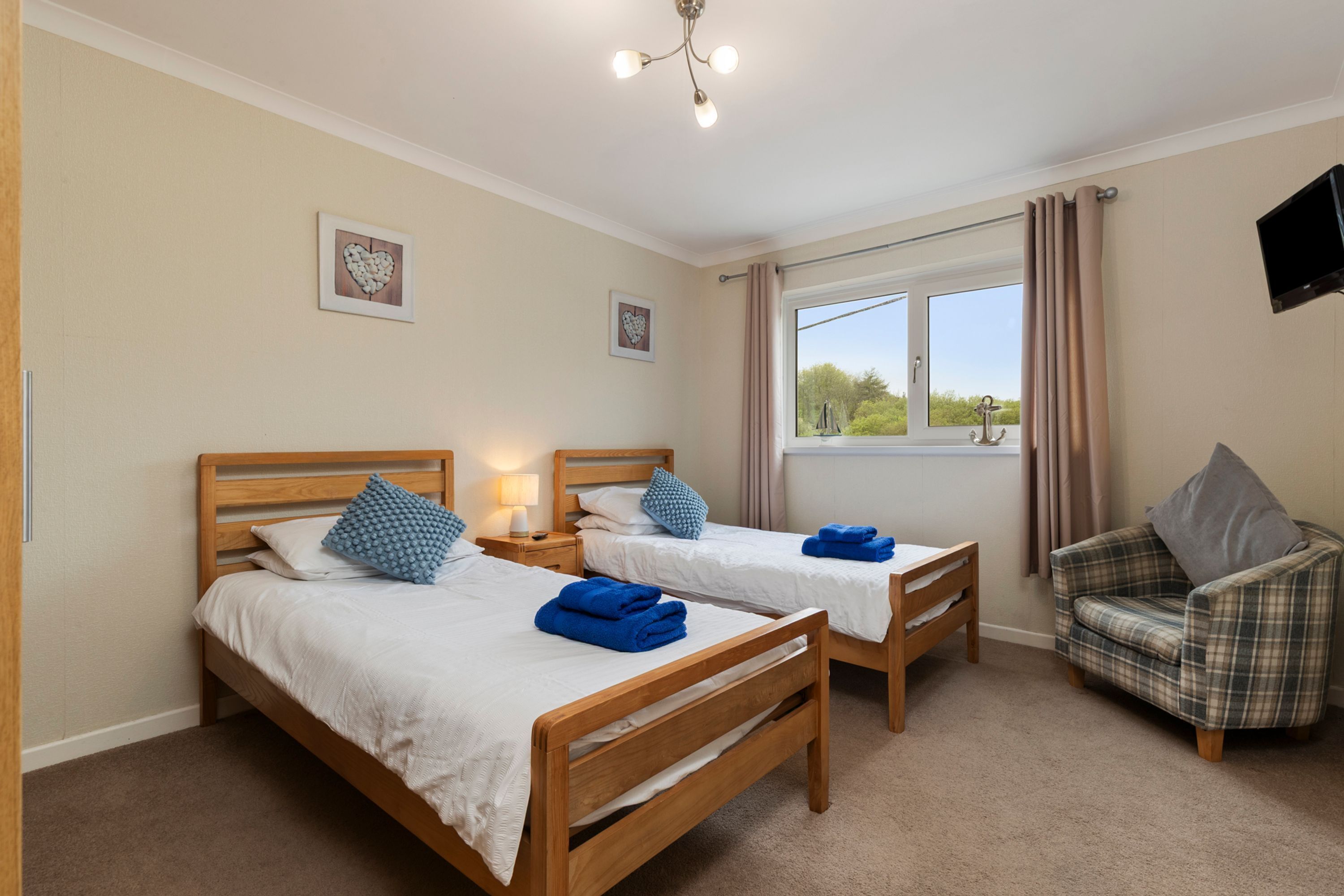 A hotel room with two wooden beds, a plaid armchair, a television, and a window with curtains.