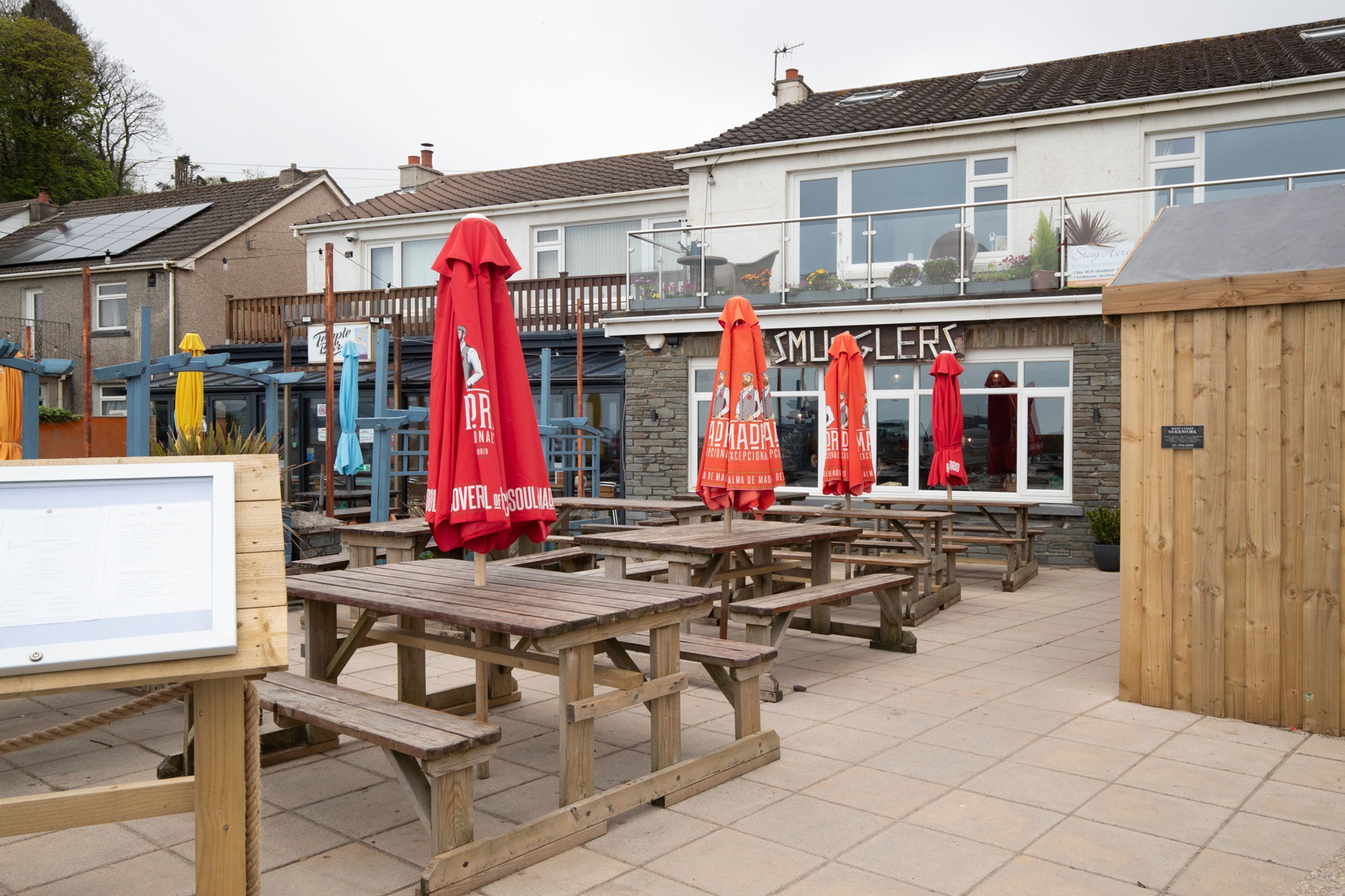 An outdoor seating area with wooden tables and red umbrellas, a stone building with a sign, and a wooden fence.
