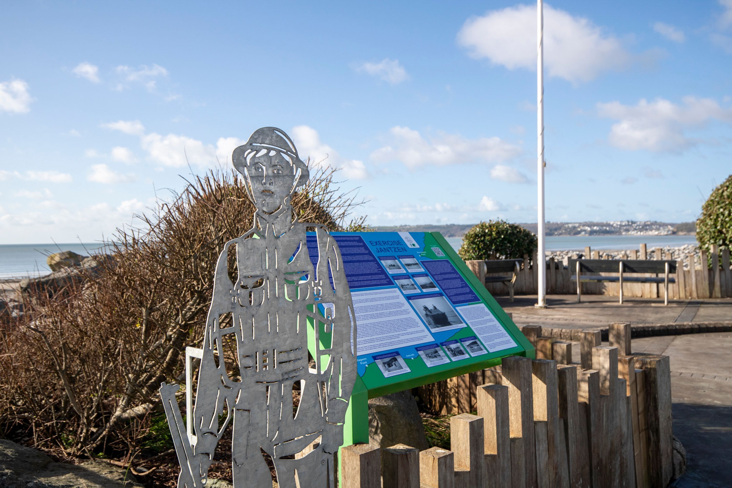 A metal sculpture with a green informational board, set against a coastal backdrop with a wooden fence and a clear sky.