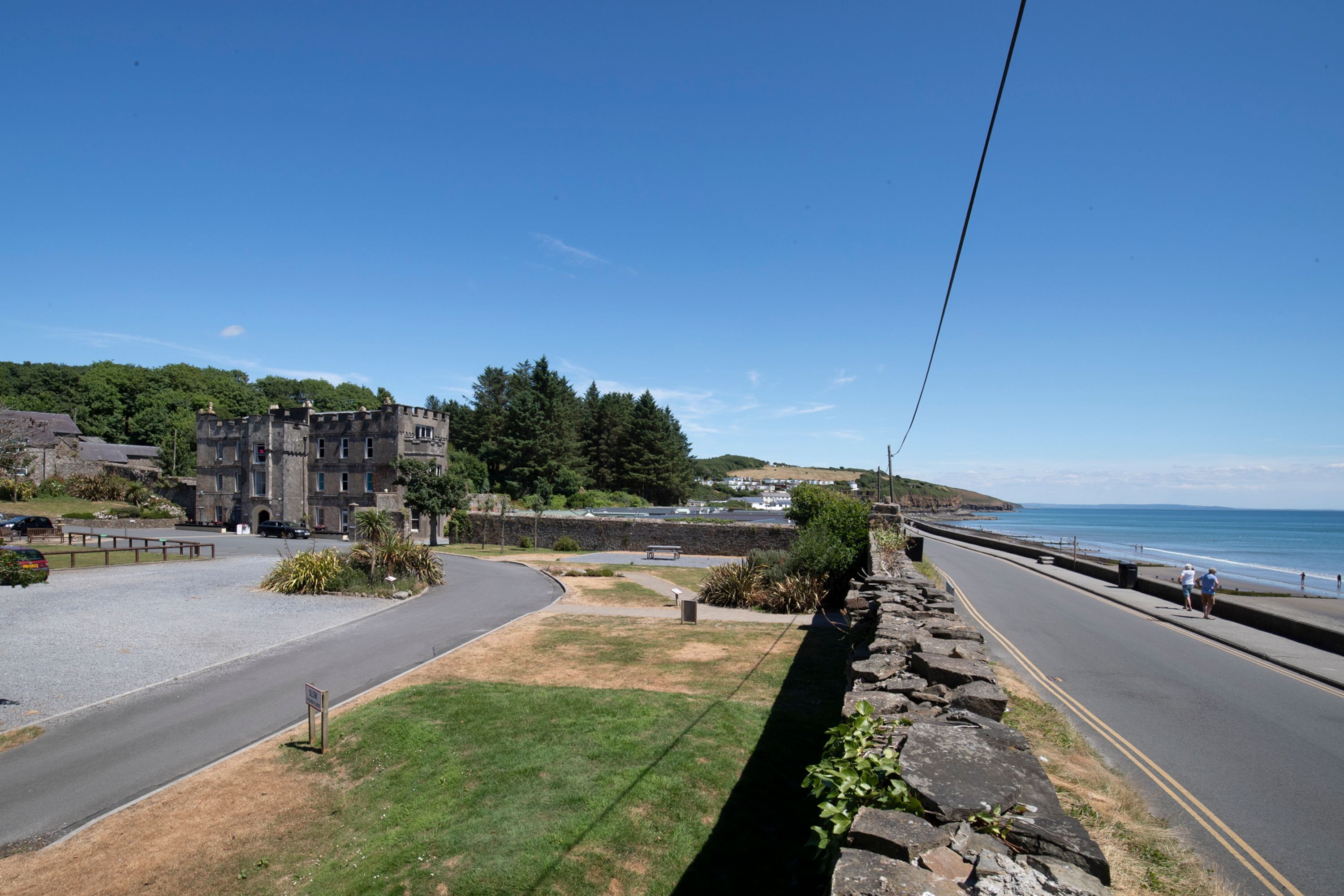 A coastal road with a stone wall, a grassy area, and a building with multiple windows.