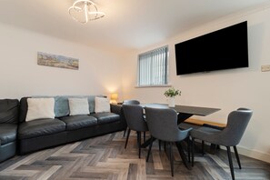 Un salon moderne comprenant un canapé, des fauteuils, une table à manger, un téléviseur à écran plat, un tableau et un parquet à motifs en chevrons.