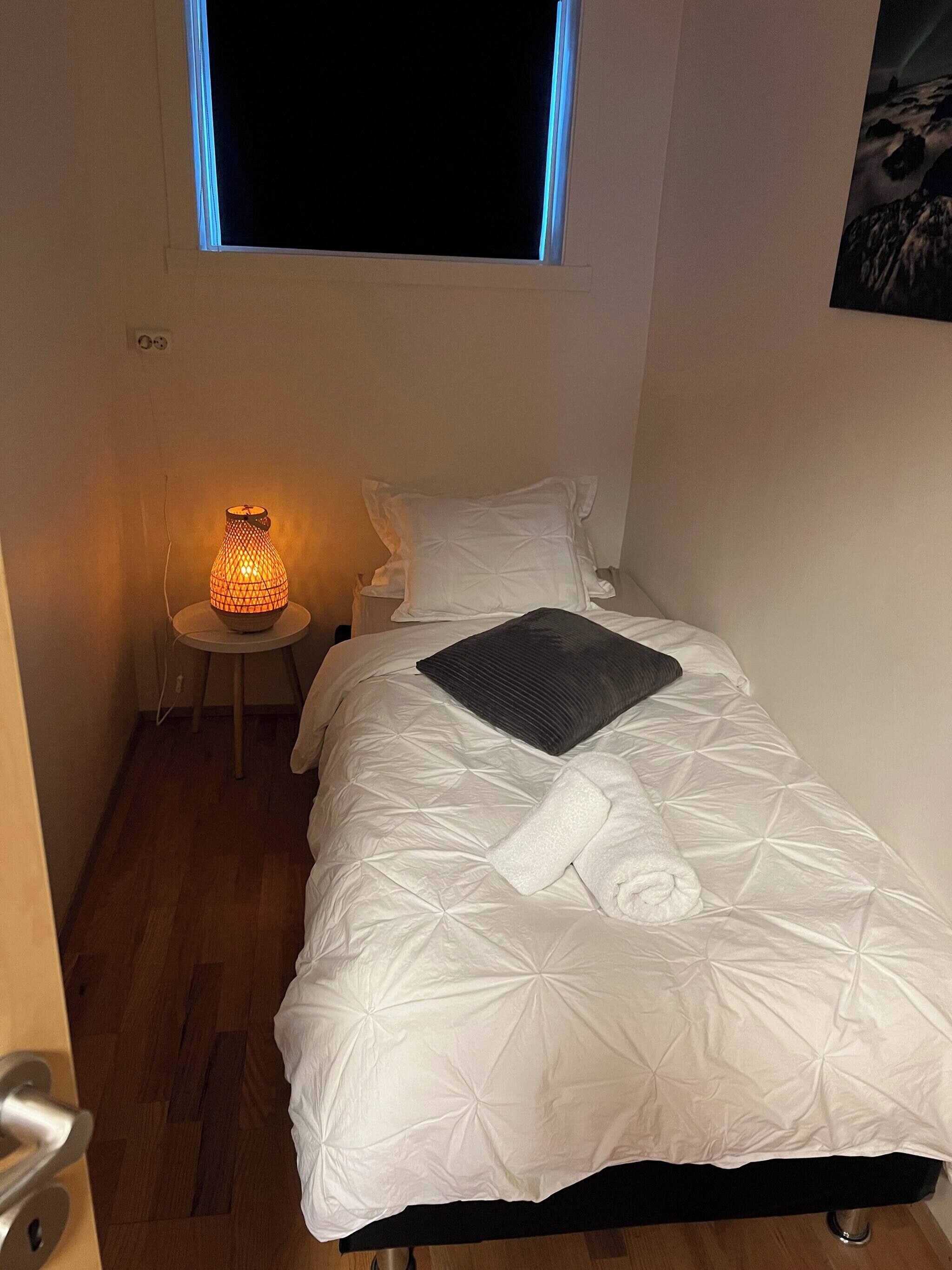 A small, neatly made bed with a white quilt, a rolled towel, and a dark pillow. There is a small table with a lamp next to the bed.
