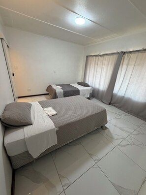 Ein Schlafzimmer mit zwei Betten, einer Deckenleuchte, einem Fenster mit Vorhängen und einem Fliesenboden.
