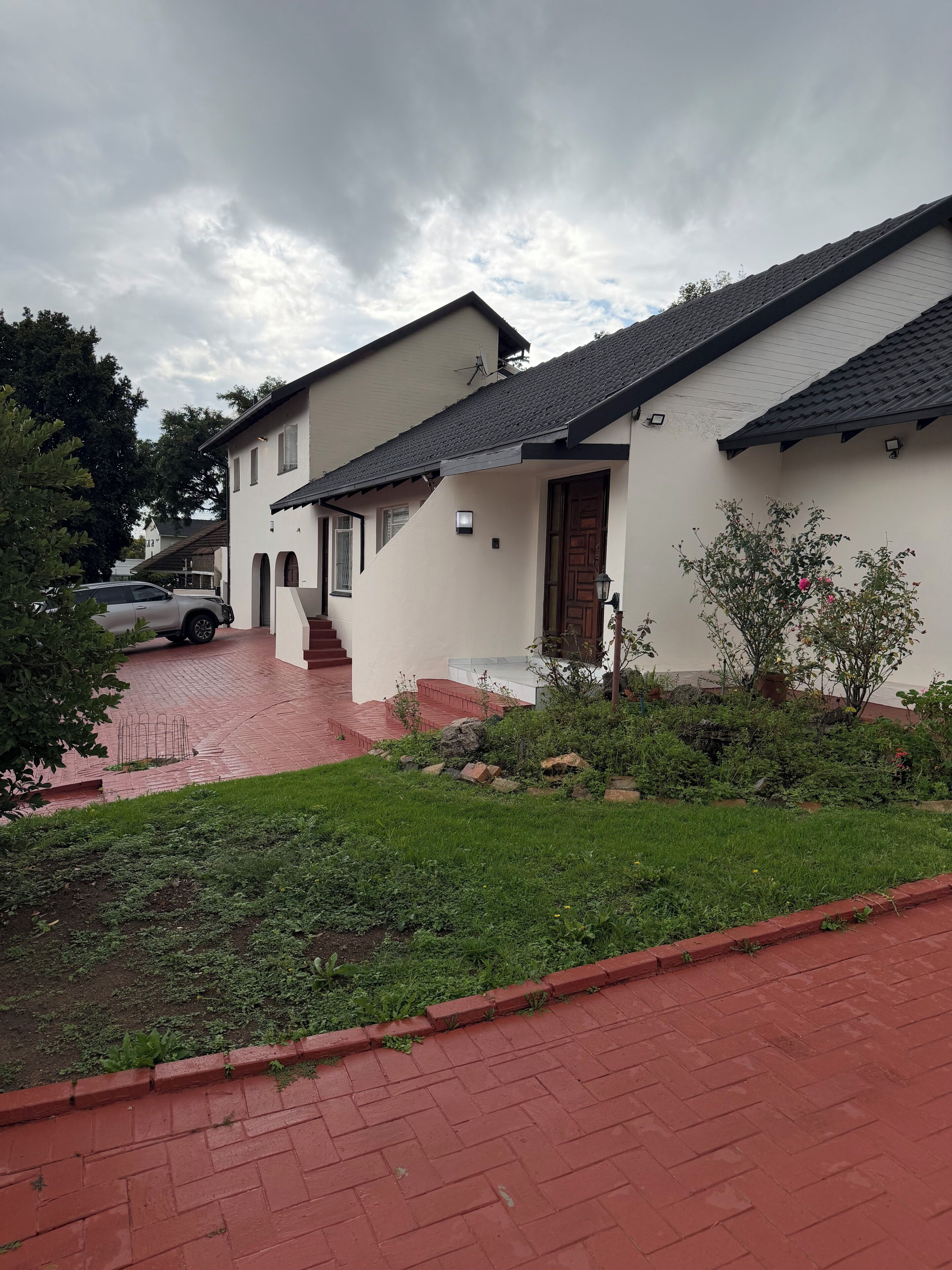 A two-story house with a red brick driveway, a small garden with plants and a tree, and a car parked in the background.