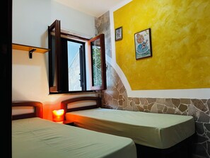 Ein Zimmer mit zwei Betten, einem Fenster mit offenem Vorhang, einer Wand mit Steinmusterung und einem gerahmten Bild.