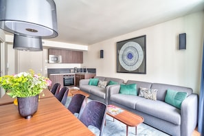 Ein modernes Wohnzimmer mit grauem Sofa, einem Esstisch mit Stühlen, einer Küchenzeile und einem großen abstrakten Gemälde an der Wand.