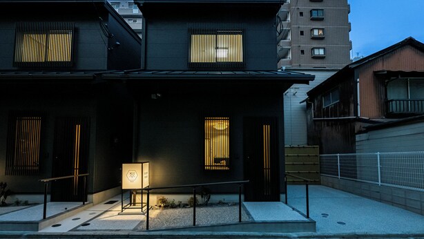 Un edificio moderno de dos pisos con fachada oscura, ventanas amplias con persianas verticales, un pequeño espacio al aire libre para sentarse y una escalera visible que conduce a la entrada.