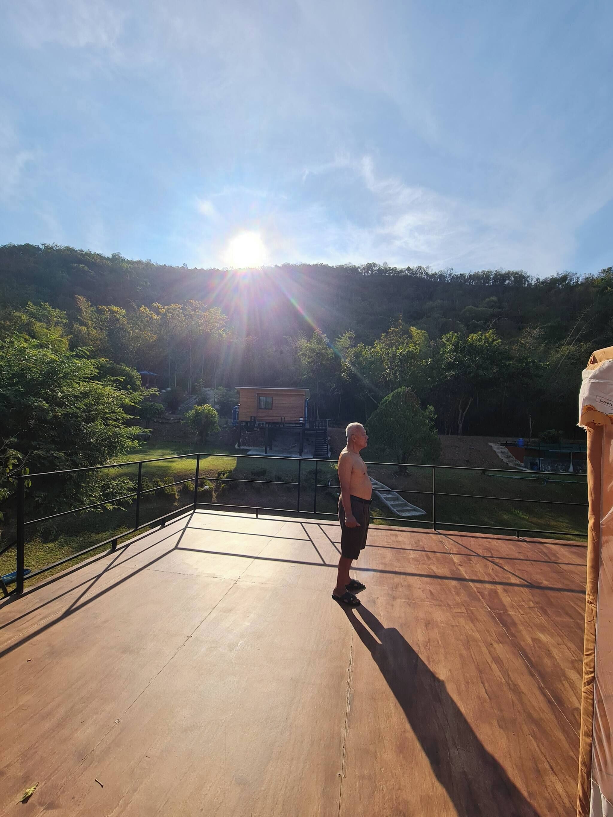 A person standing on a wooden deck with a view of a forested hillside and a building in the background.