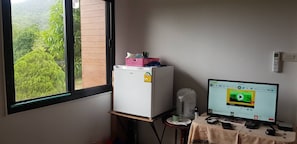 Une pièce avec une fenêtre, un réfrigérateur blanc, une petite table avec un ordinateur, et une vue sur la verdure à l’extérieur.