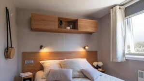 A bedroom with a wooden headboard, bedside lamps, a nightstand, and a window with curtains. - Pocket" mobile home in the heart of the pony club (Hostens)