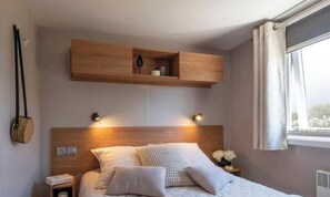 A bedroom with a wooden headboard, bedside lamps, a nightstand, and a window with curtains. - Pocket" mobile home in the heart of the pony club (Hostens)