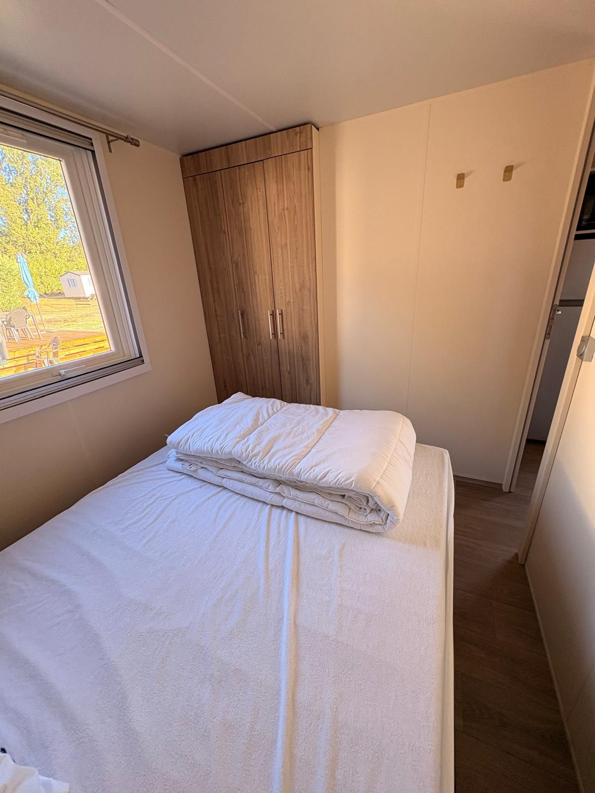 Una cama pequeña y bien hecha con ropa blanca en una habitación compacta, con un armario de madera y una ventana.