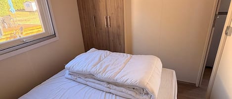 Una cama pequeña y bien hecha con ropa blanca en una habitación compacta, con un armario de madera y una ventana.