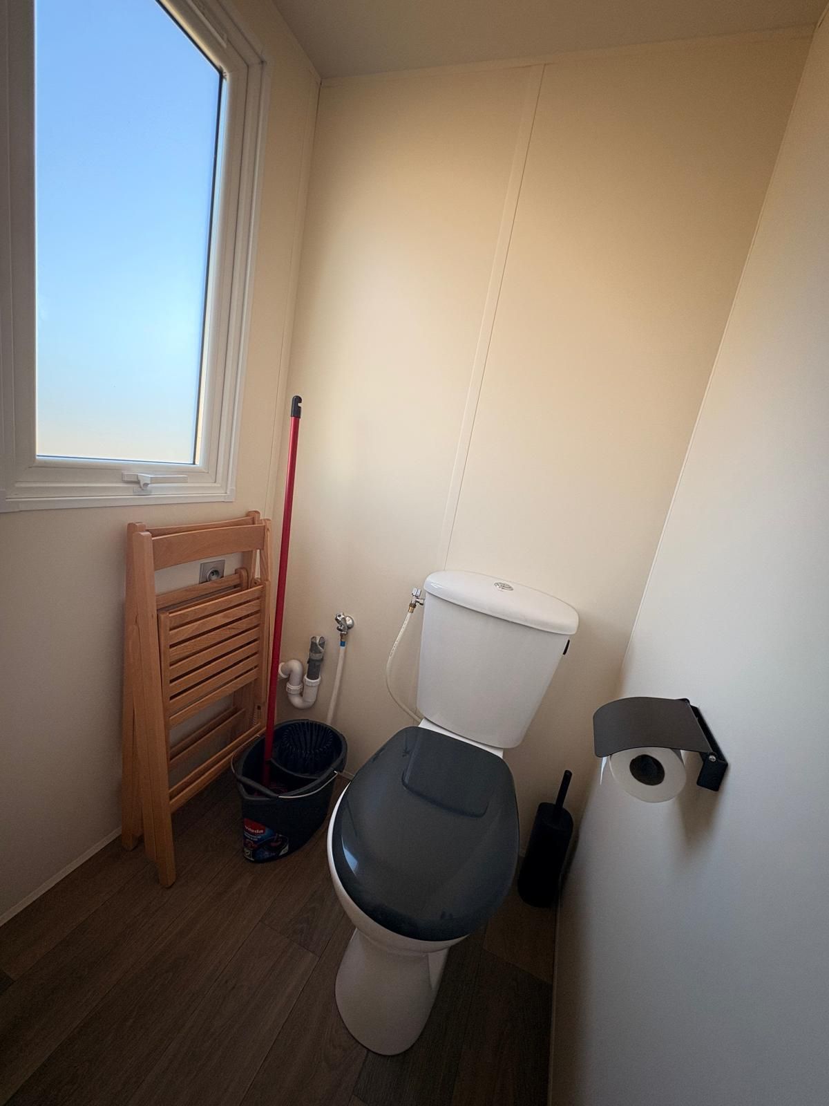 Een kleine badkamer met toilet, een houten rek, een zwarte prullenbak en een raam.