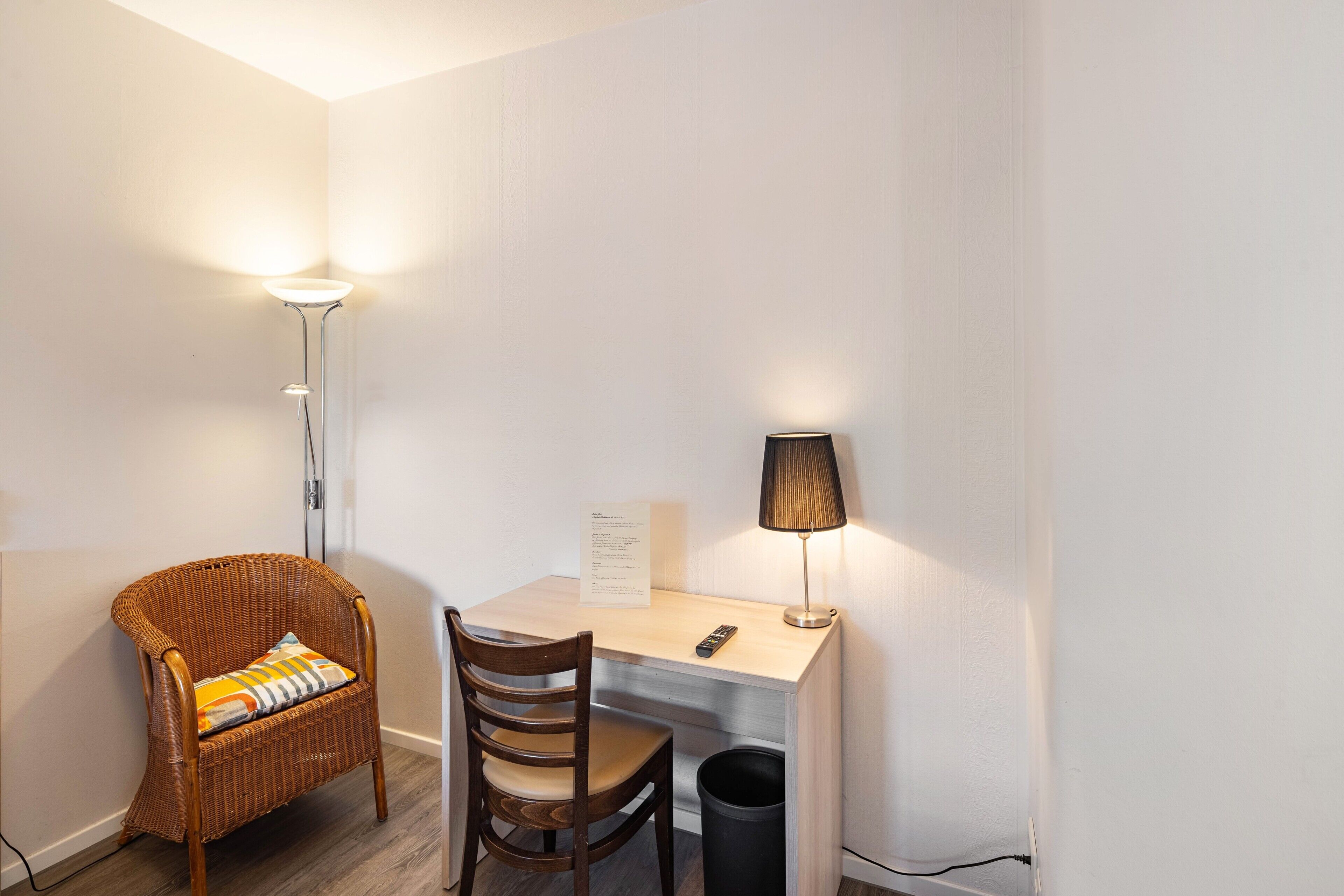 Ein kleines, sauberes Zimmer mit Schreibtisch, Korbstuhl, Lampe und Papierkorb.