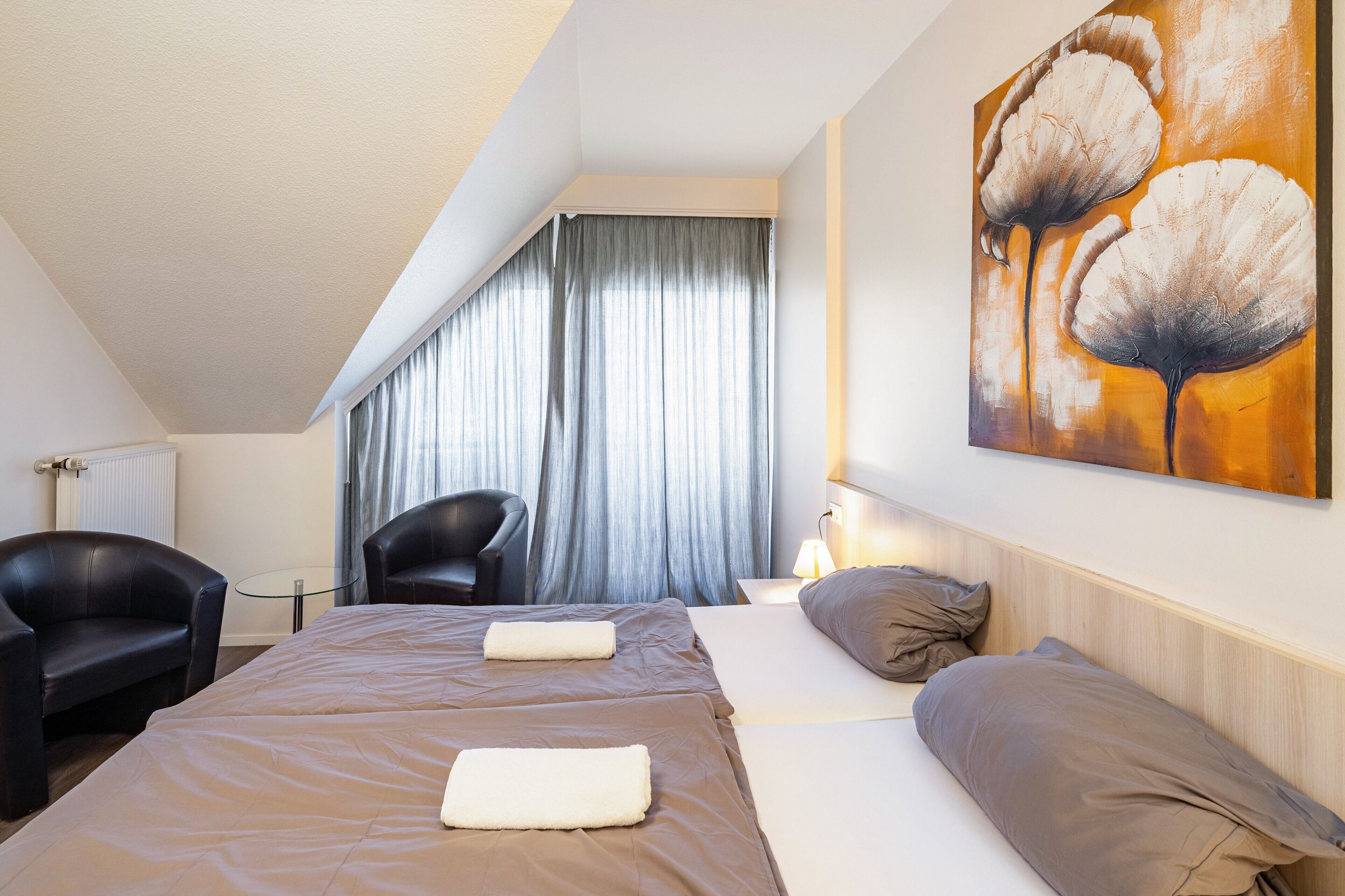 A hotel room with a bed, two chairs, a painting, and a window with curtains.