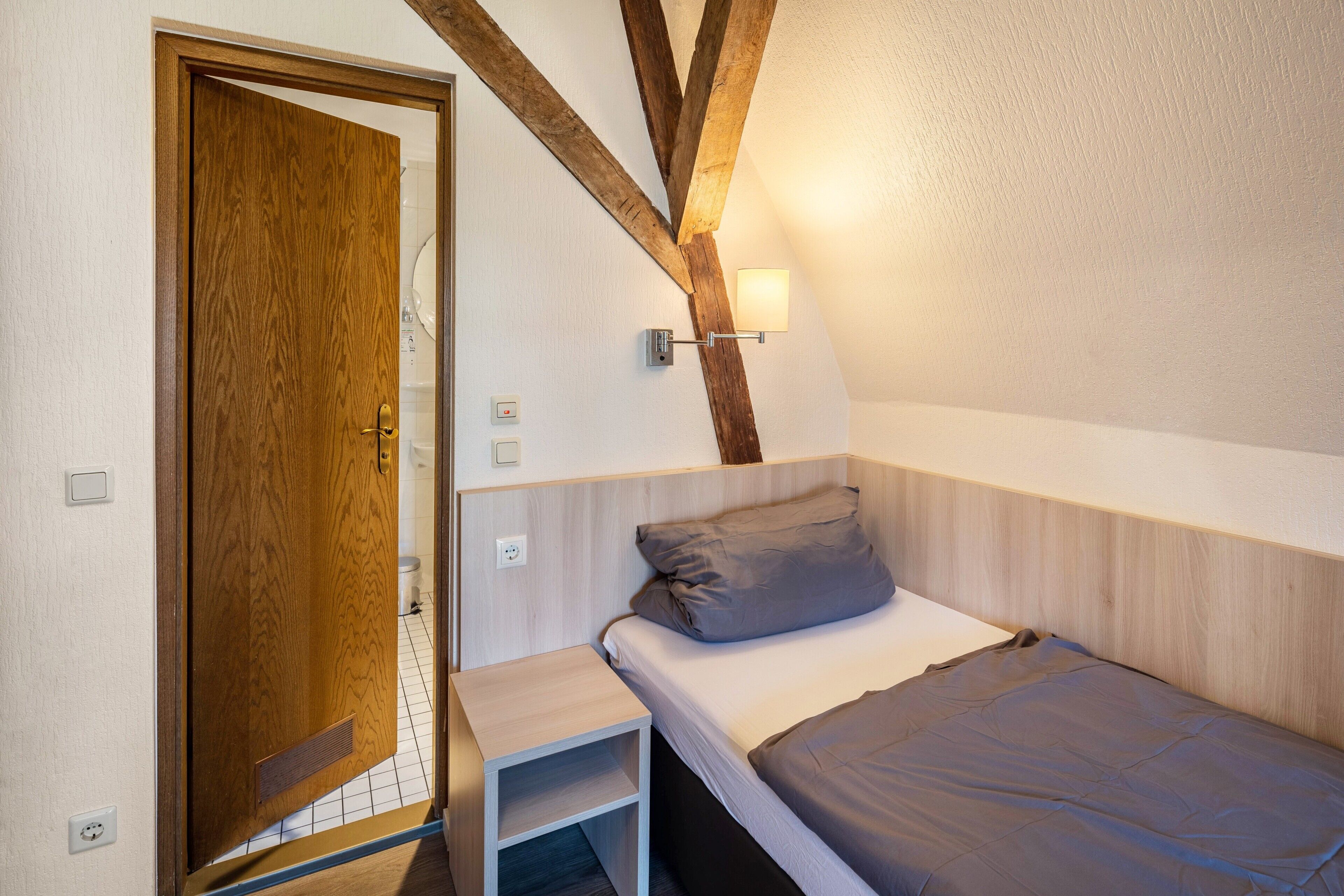 Ein Schlafzimmer mit Holztür, einem Bett mit grauer Matratze, einem kleinen Nachttisch und einer Wandleuchte.