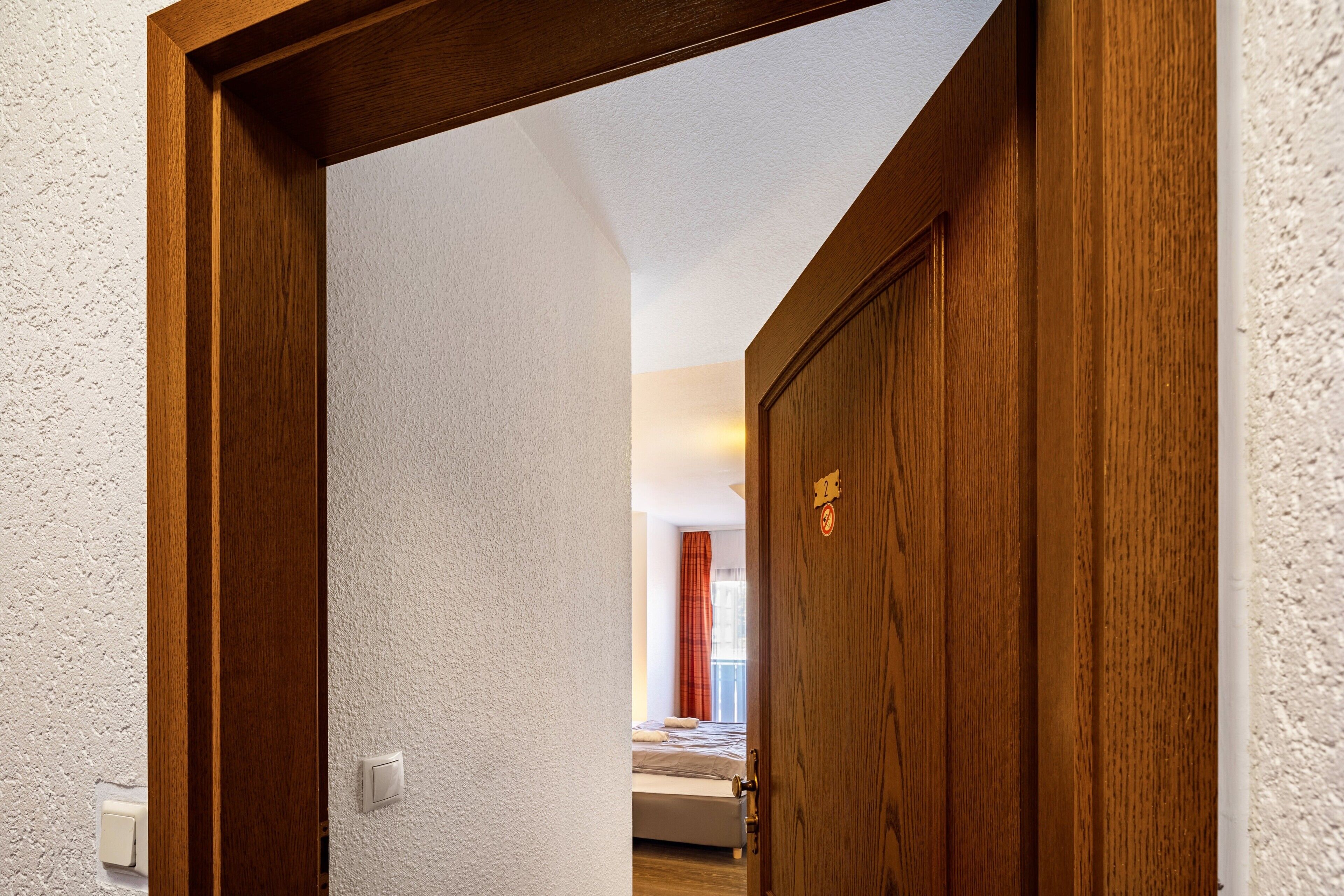 Una porta de fusta que dona a un dormitori amb un llit, una finestra amb cortines i un interruptor a la paret.