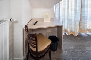 Een kleine, verzorgde kamer met een houten bureau, een stoel, een zwarte prullenbak en een radiator.