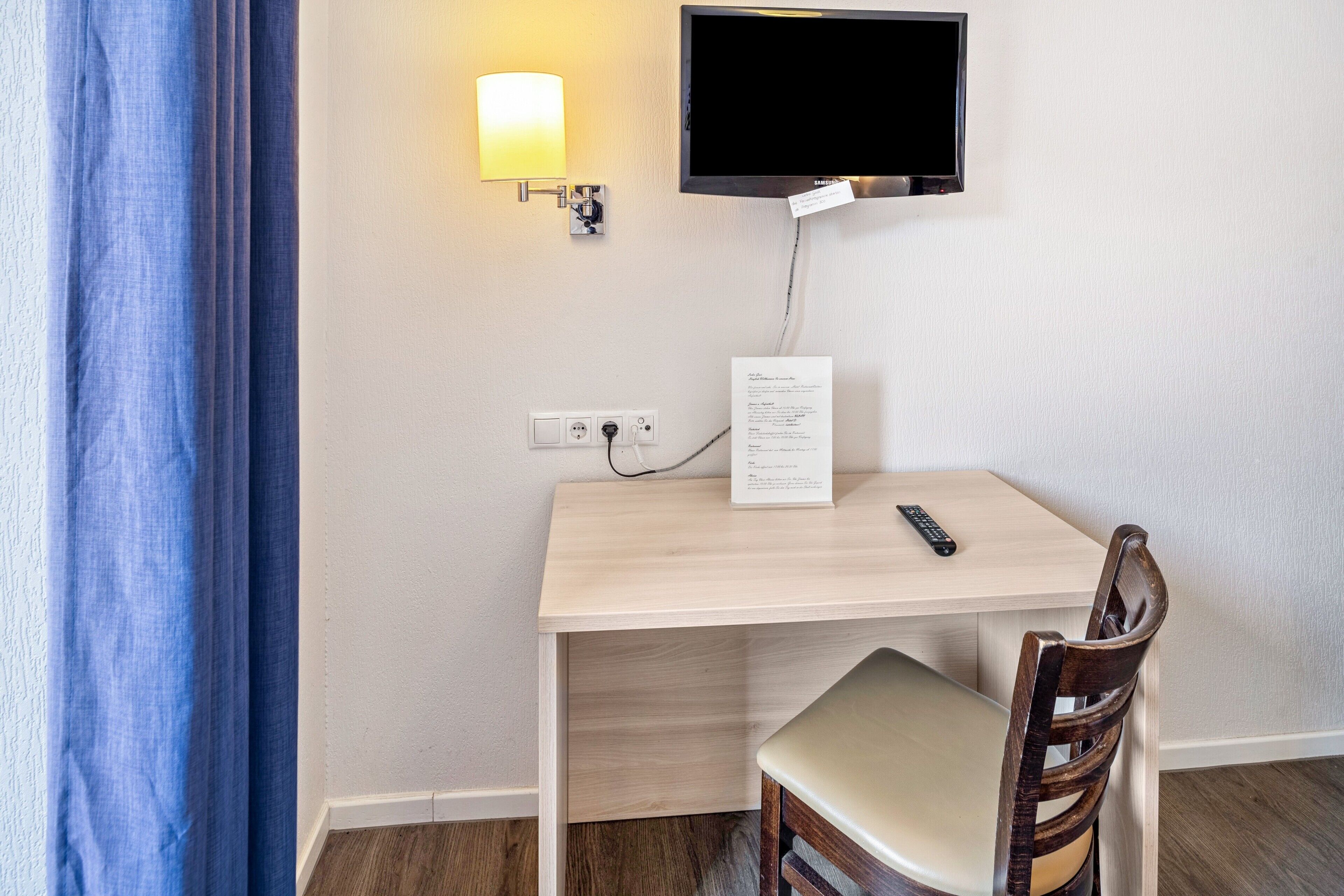 A hotel room with a desk, chair, TV, lamp, and a sign on the wall.