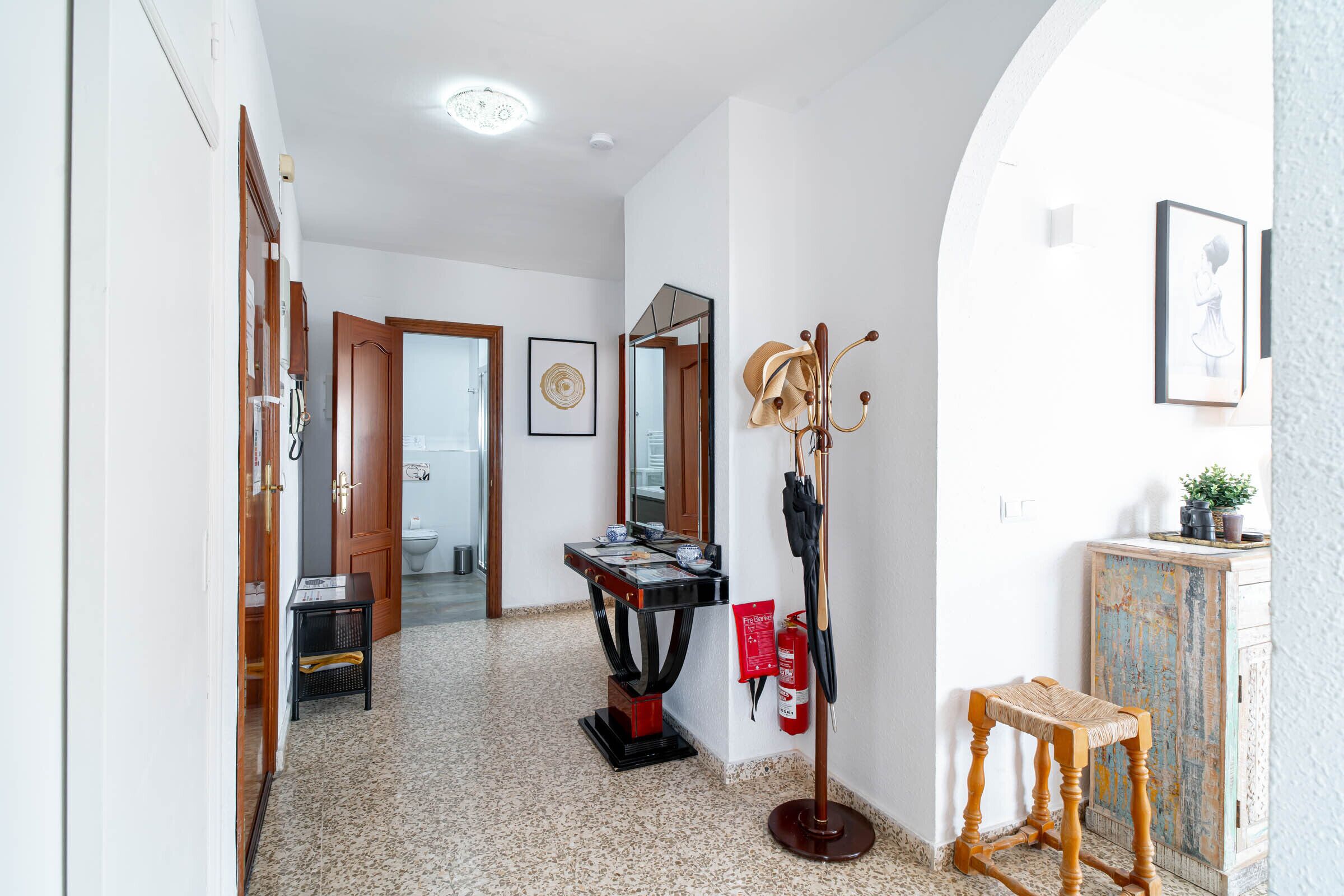 Un pasillo con un banco de madera, perchero, extintor, espejo y puerta que da a un baño.