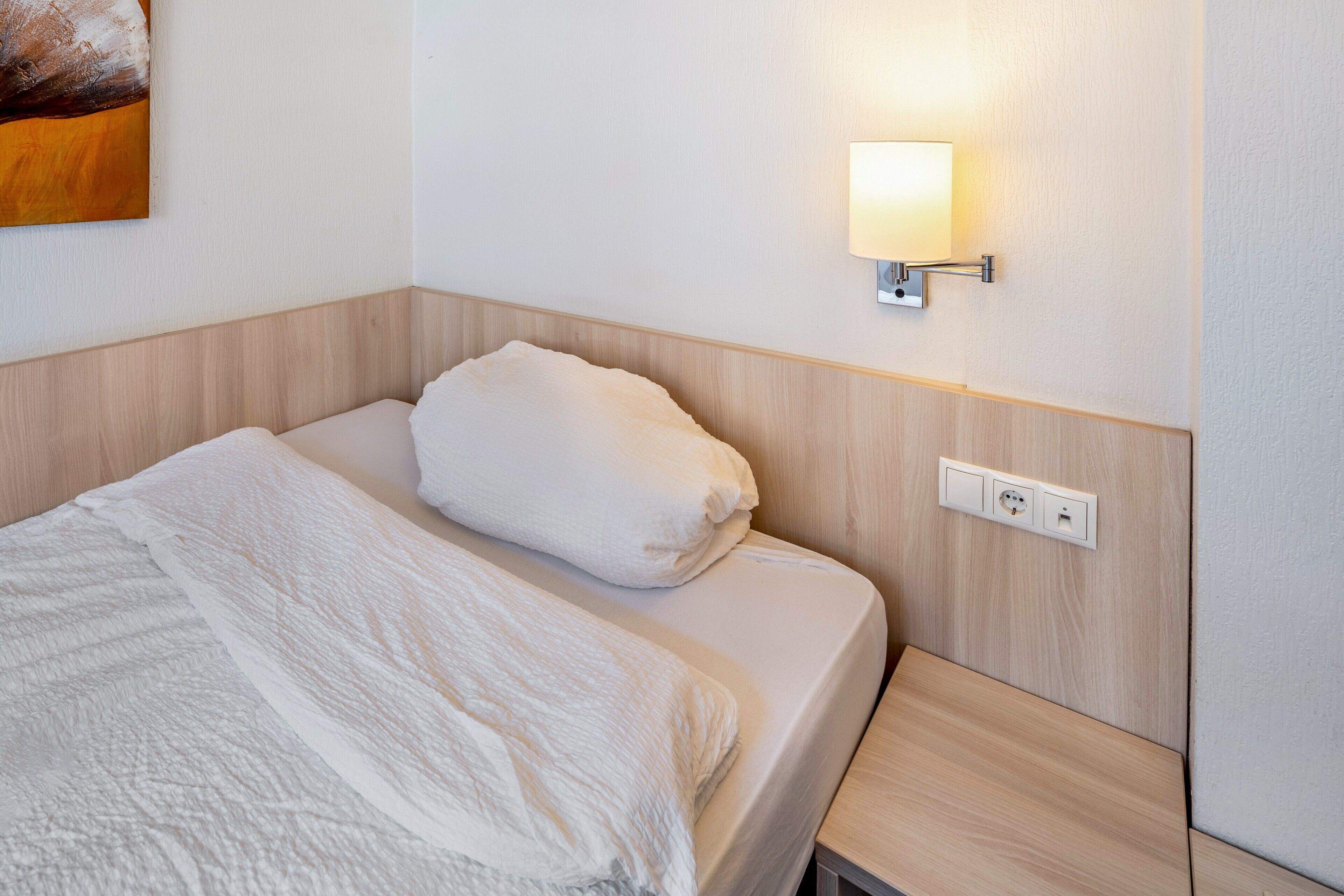 En pænt redt seng med hvid dyne og pude, et natbord med lampe og en stikkontakt.
