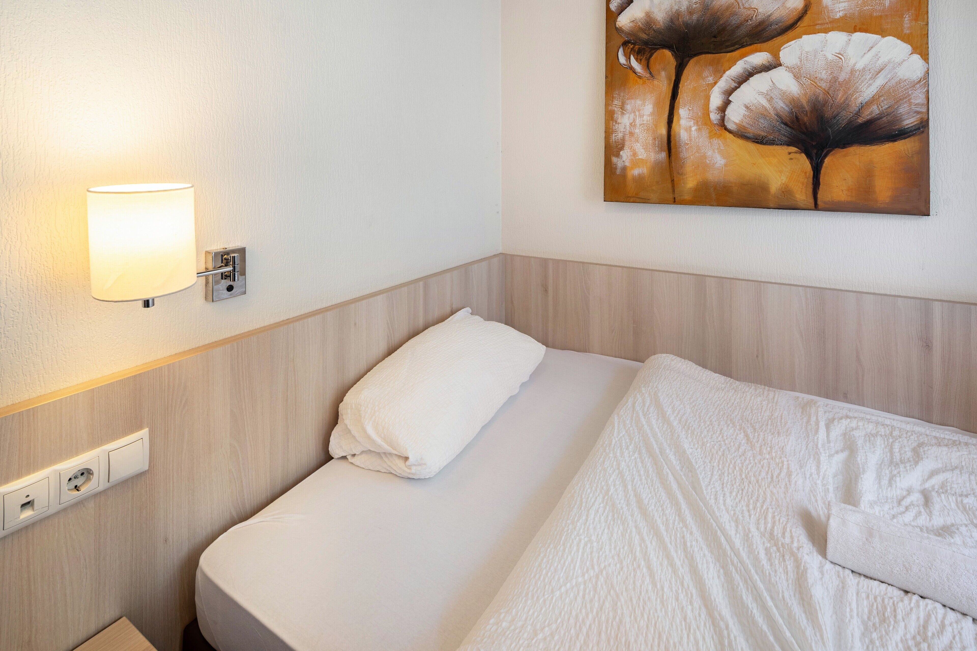 A hotel room with a bed, a wall-mounted lamp, a painting, and a wooden headboard.