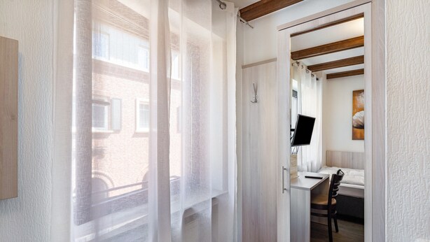 Ein Zimmer mit einem Bett, einem Schreibtisch, einem Stuhl und einem Fenster mit durchsichtigen Vorhängen.