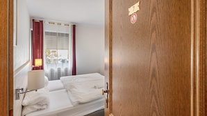 A hotel room with a bed, a bedside table with a lamp, a window with curtains, and a door with a number sign.