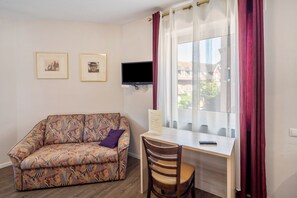 Ein Zimmer mit einer Couch, einem Schreibtisch mit Stuhl, einem an der Wand befestigten Fernseher und einem Fenster mit Vorhängen.