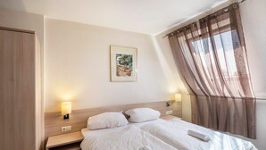 A hotel room with two beds, a nightstand, a wall-mounted lamp, a framed picture, and a window with curtains. - Zimmer „Doppelzimmer 14“ mit WLAN (Rhede)