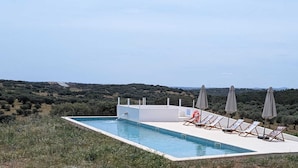 Une piscine rectangulaire dotée d’une terrasse blanche, entourée de chaises longues et de parasols, le tout se fondant dans un paysage d’oliviers et de collines.
