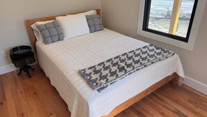 Une chambre avec un lit en bois, une literie blanche, un plaid à motifs, une petite table de chevet noire et une fenêtre donnant sur l’extérieur.