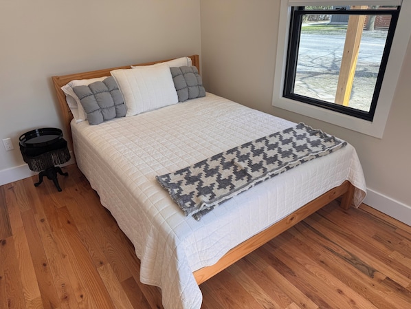 A bedroom with a wooden bed, white bedding, a patterned throw, a small black side table, and a window with a view of the outdoors.