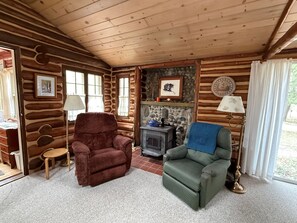 L’intérieur d’une cabane en rondins, avec une cheminée, deux fauteuils, une lampe et un mur en pierre.