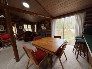 L’intérieur d’une cabane en rondins, avec une table à manger en bois, des chaises à revêtement rouge, une cheminée et une grande fenêtre avec des rideaux.