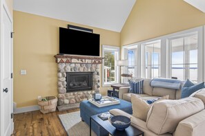Una sala de estar con chimenea de piedra, un televisor montado encima, un sofá color beige con cojines azules y vistas amplias a través de ventanas grandes.