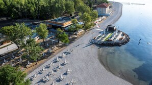 Vista aérea de una propiedad frente a la playa con una fila de sillas y sombrillas, un edificio de techo rojo y un muelle que se adentra en el agua.