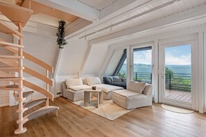 Ein stilvolles Loft mit einer Wendeltreppe aus Holz, einer Sofagarnitur, einem kleinen Tisch und großen Schiebeglasfenstern, die auf einen Balkon mit Blick eröffnen.