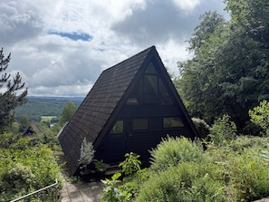 Eine dreieckige Holzhütte mit steiler Dachneigung, umgeben von dichtem Grün und einem schönen Ausblick.