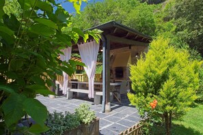 A covered outdoor seating area with a wooden table and chairs, surrounded by greenery and a small tree with a red flower.