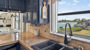 Uma cozinha com armários azul escuro, uma janela com vista para a água e edifícios, uma pia com torneira moderna e um escorredor de louça.