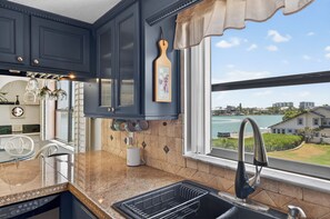 A kitchen with dark blue cabinets, a window with a view of a body of water and buildings, a sink with a modern faucet, and a dish rack. - 2BR 2Ba Treasure Island Luxury waterfront updated Condo., steps to beach access (Treasure Island)