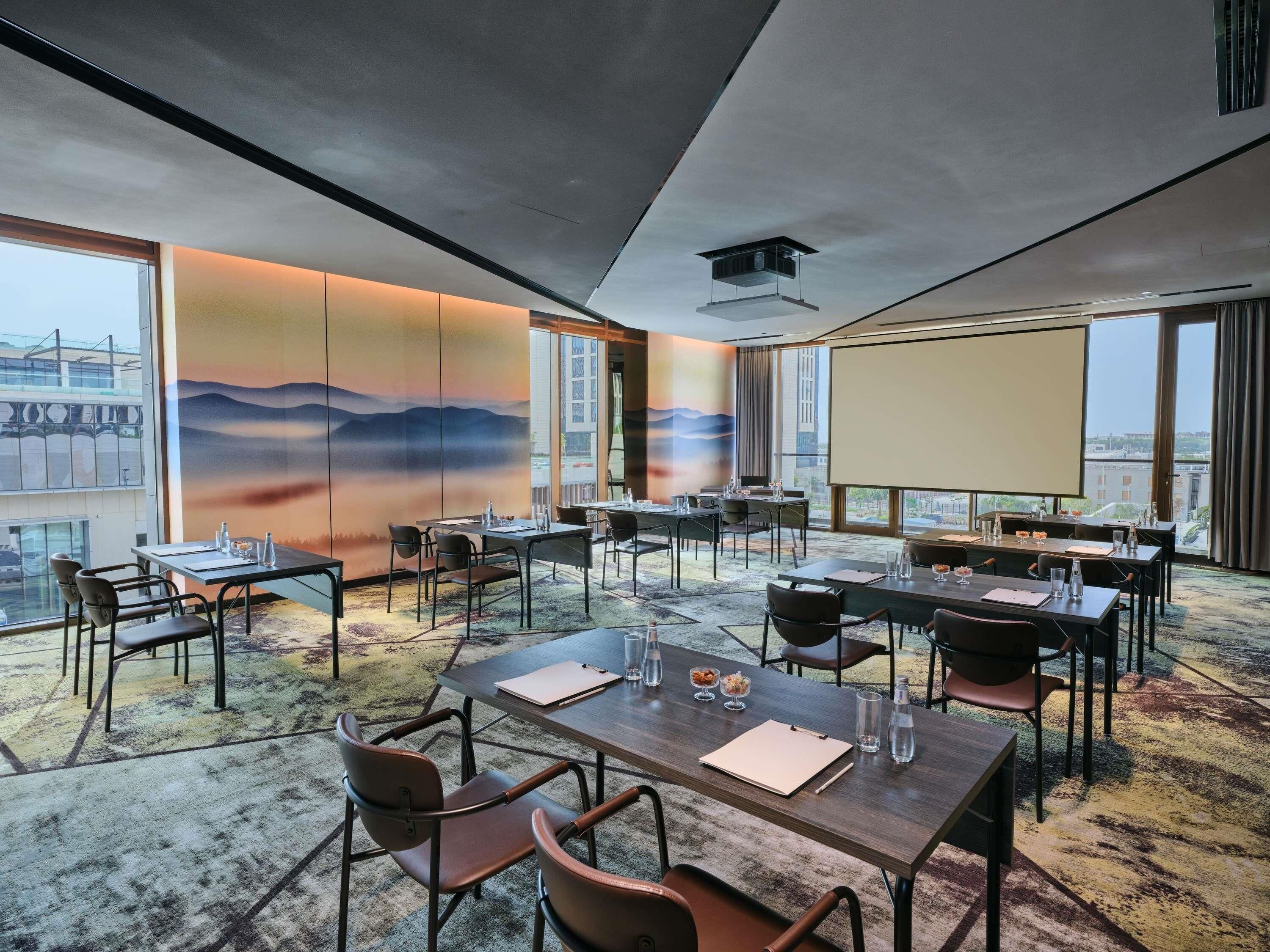 A conference room with tables, chairs, a projector screen, and a large window with a city view.