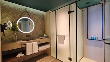 A modern bathroom with a herringbone wood wall, a round mirror with a light, a glass shower enclosure, and a wooden vanity with towels.