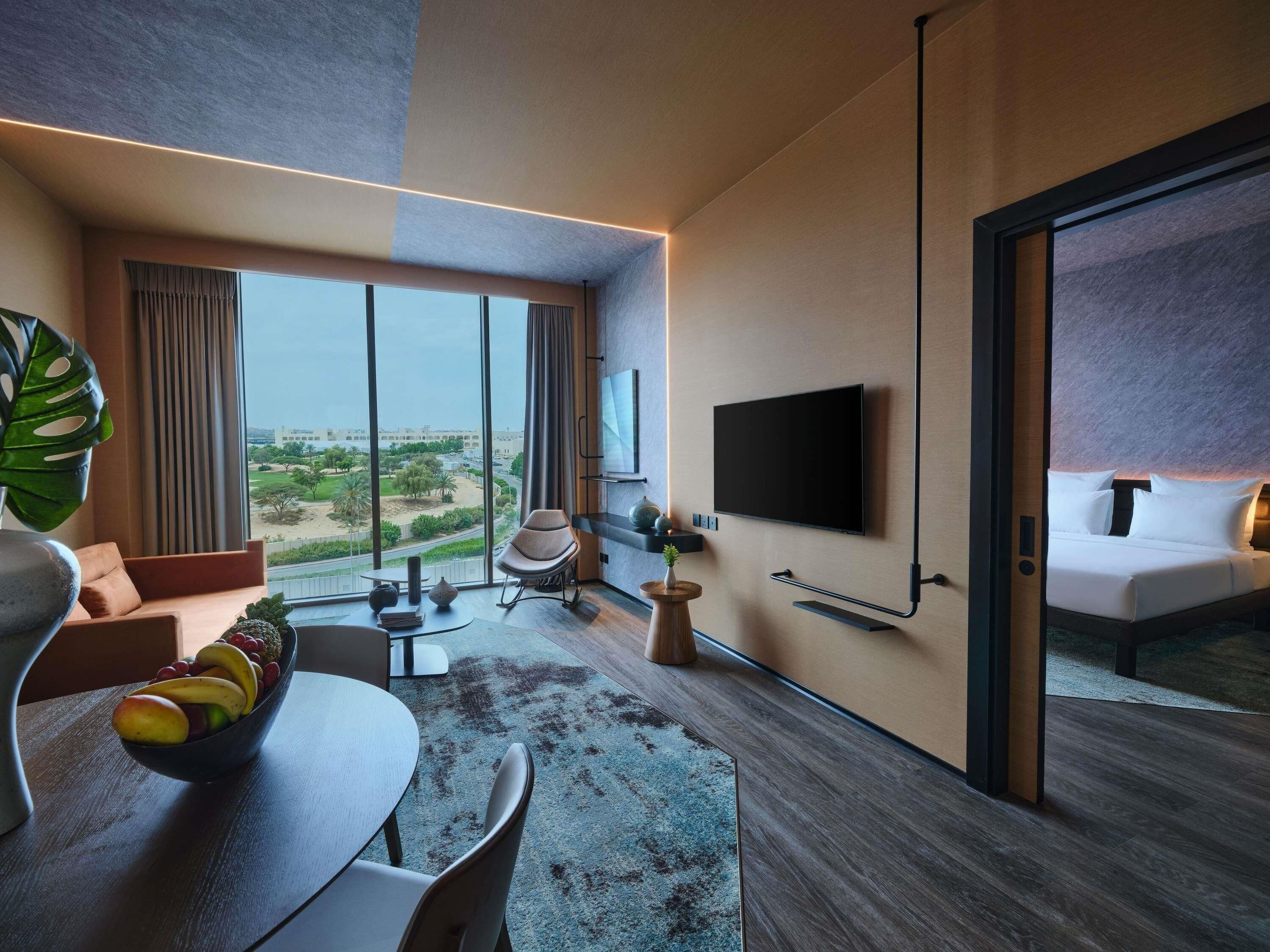A modern hotel room with a large window, a flat-screen TV mounted on the wall, a dining area with a table and chairs, and a bed with a headboard.